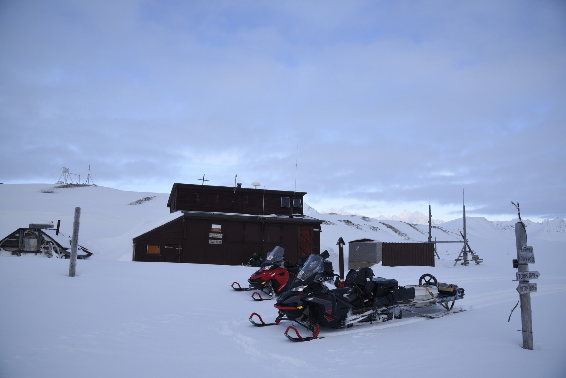 Na pierwszym planie fotografii skutery śnieżne, dalej zabudowania stacji badawczej, a w tle góry. Całość pokryta śniegiem.