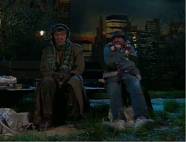 Zdjęcie przedstawia dwóch mężczyzn siedzących na ławce w parku. Obaj są ubrani w zniszczone rzeczy. Mężczyzna po lewej stronie jest starszy. Ma pociągłą twarz z wyraźnie zaznaczonymi zmarszczkami. Oczy głęboko osadzone, opuchnięte. Głowę i szyję ma owiniętą szalem. Na dłoniach ma rękawiczki bez palców. Ubrany jest w długi płaszcz. Mężczyzna siedzi ze spuszczonymi oczami. Po prawej stronie znajduje się dojrzały mężczyzna o owalnej twarzy, ze szpakowatym zarostem. Na głowie ma kapelusz, Ubrany jest w kurtkę, spodnie, pod szyją ma zawinięty szalik, Trzyma nogi wyciągnięte przed siebie. Patrzy się na siedzącego obok mężczyznę. Za plecami mężczyzn znajdują się zabudowania miejskie: domy, budynki, wieżowce. Jest ciemno, palą się latarnie.