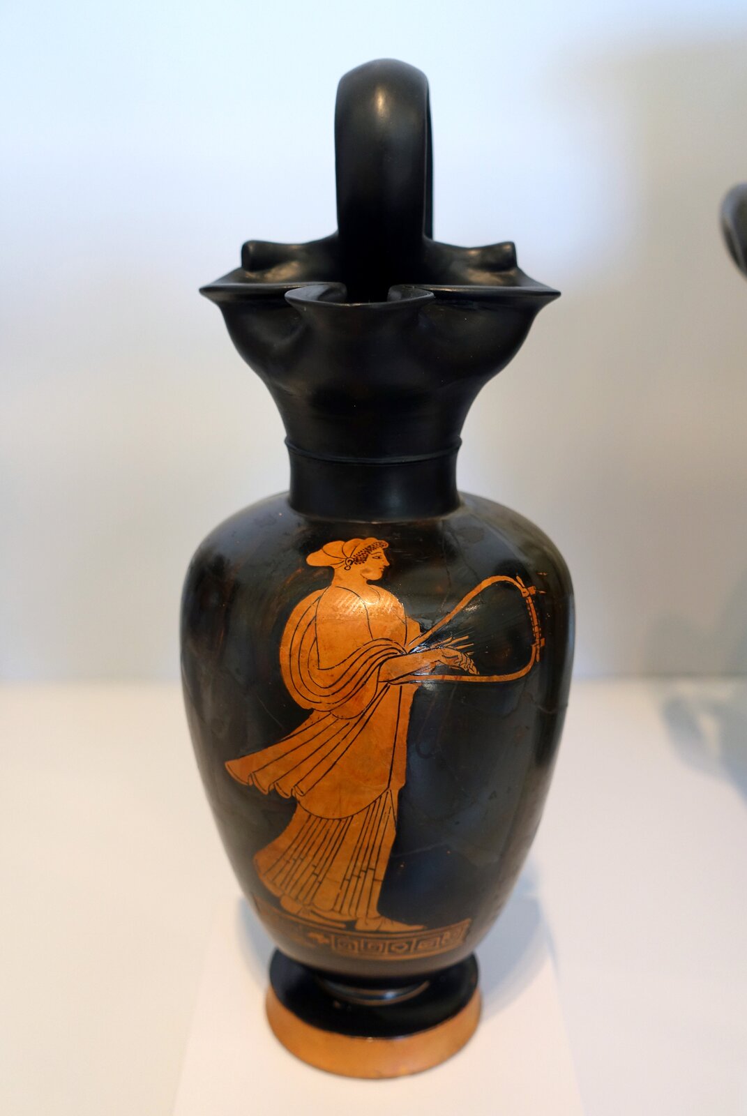 Ilustracja przedstawia naczynie antyczne. Jest to flakon wykonany w stylu czerwonofigurowym. Jego górna część ma kształt płatków kwiatu. Naczynie jest czarne, malowidło zaś pomarańczowe. Na brzuścu przedstawiona została postać młodej kobiety o włosach spiętych w kok i ukrytych pod welonem. Ubrana jest w długą suknię i gra na harfie.