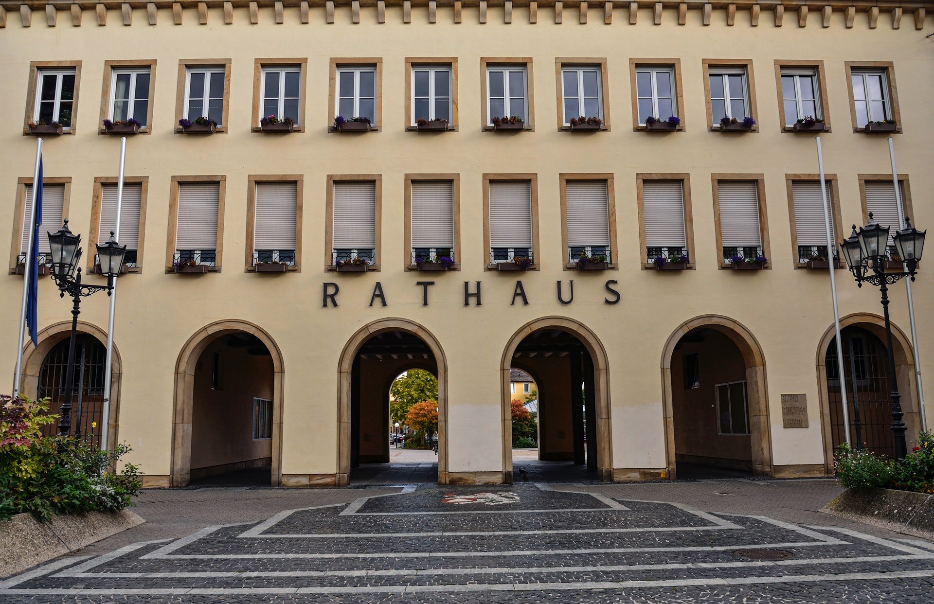 Zdjęcie przedstawia budynek urzędowy z jasną elewacją. Ma dwa piętra, a na parterze rząd łuków. Na parapetach okien są kwiaty. Nad wejściem jest napis: Rathaus. 