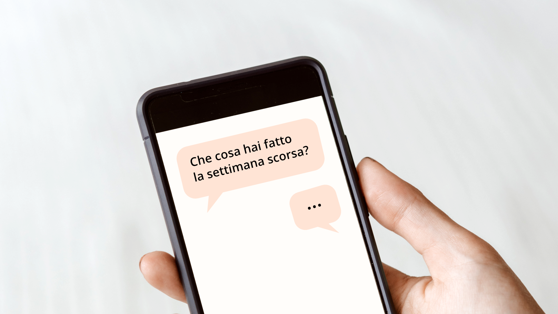 Zdjęcie przedstawia telefon komórkowy, który znajduje się w dłoni. Na ekranie telefonu widoczny jest czat. W pierwszym dymku czatu znajduje się tekst: "Che cosa hai fatto la settimana scorsa?". W drugim dymku znajdują się trzy kropki.