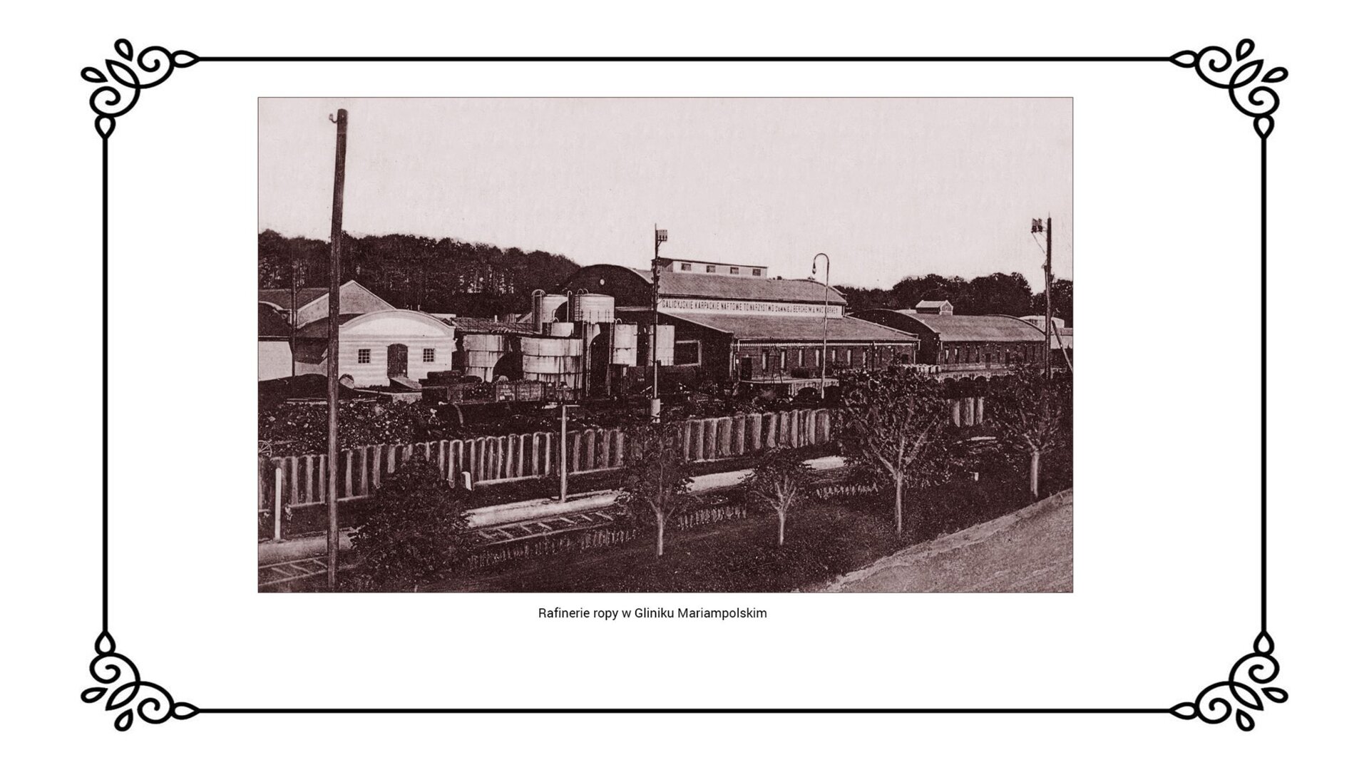 Slajd przedstawia krajobraz industrialny Glinika Mariampolskiego z zabudowaniami rafinerii. W centrum ilustracji widoczne są zbiorniki na przetworzoną ropę. Po lewej i po prawej stronie zdjęcia zlokalizowane są zakłady przetwórcze, w tym destylarnie. Przed nimi znajdują się tory kolejowe. 