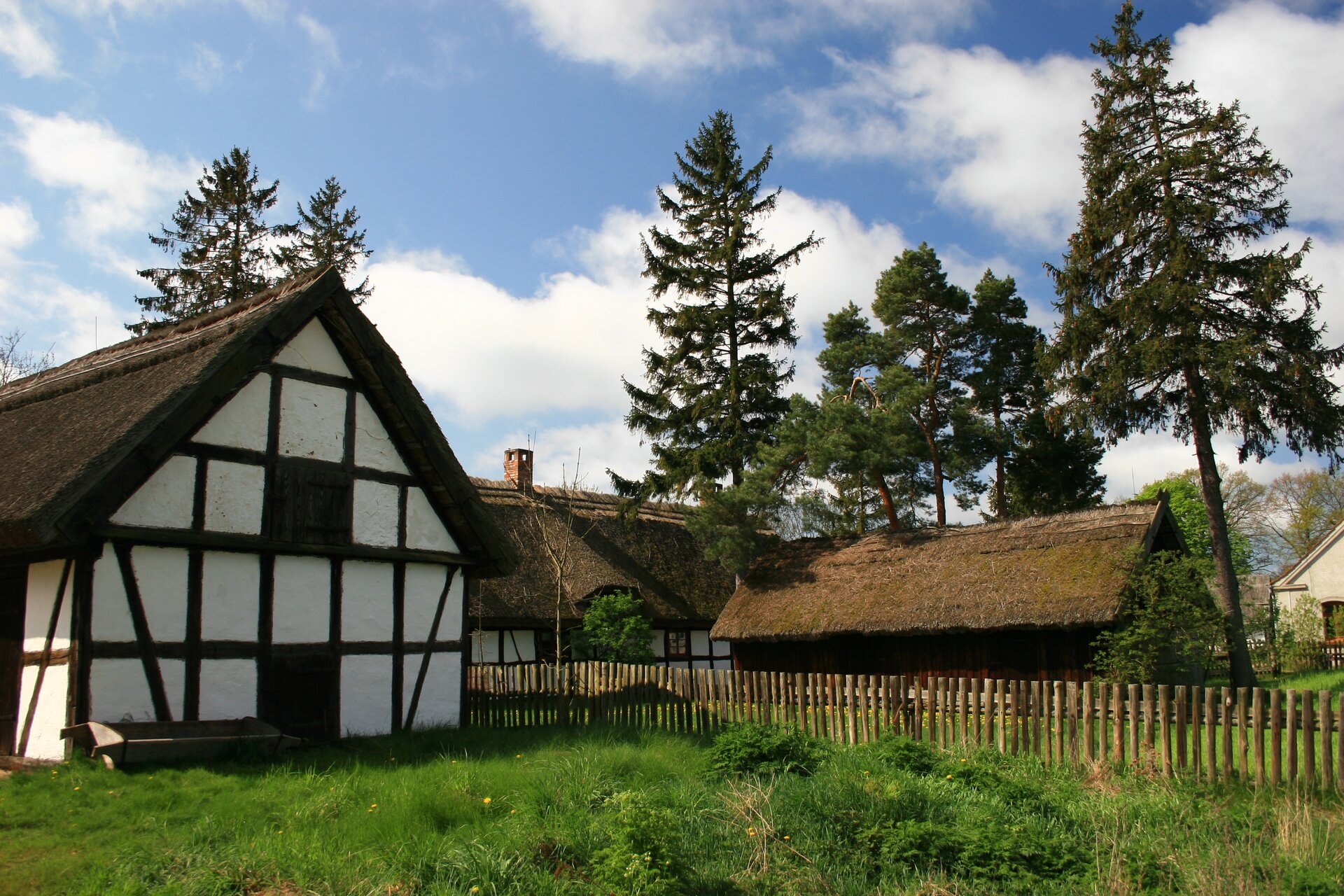 Zdjęcie przedstawia zagrodę w której znajduje się dom i budynki gospodarcze. Pierwszy budynek wysunięty na główny plan po lewej stronie jest koloru białego, na którym są umieszczone drewniane belki w kolorze ciemnego brązu, tworzą one kształt kratki. Przed budynkiem jest zielony trawnik, natomiast od prawej strony budynku wychodzi drewniany płot, za którym znajdują się pozostałe budynki. 