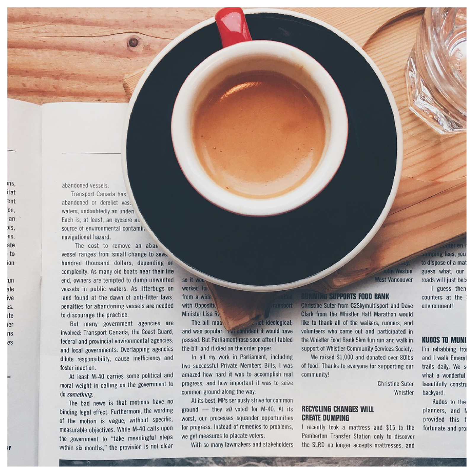 Na ilustracji znajduje się gazeta rozłożona na biurku. Na niej postawiono drewnianą deskę z filiżanką kawy oraz szklanką wody.