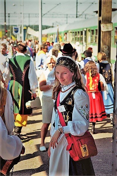 Zdjęcie przedstawia młodą dziewczynę stojącą na peronie, która jest ubrana w tradycyjny strój kaszubski. Na głowie ma szeroką czarną przepaskę haftowaną w złote wzory kwiatowe; ma również strojną, białą bluzkę z bufkami na rękawach, na których są wyhafrowane kwiaty. Posiada czarną, haftowaną w złote wzory kamizelkę, czarną spódnicę a z przodu długą, białą zapaskę.