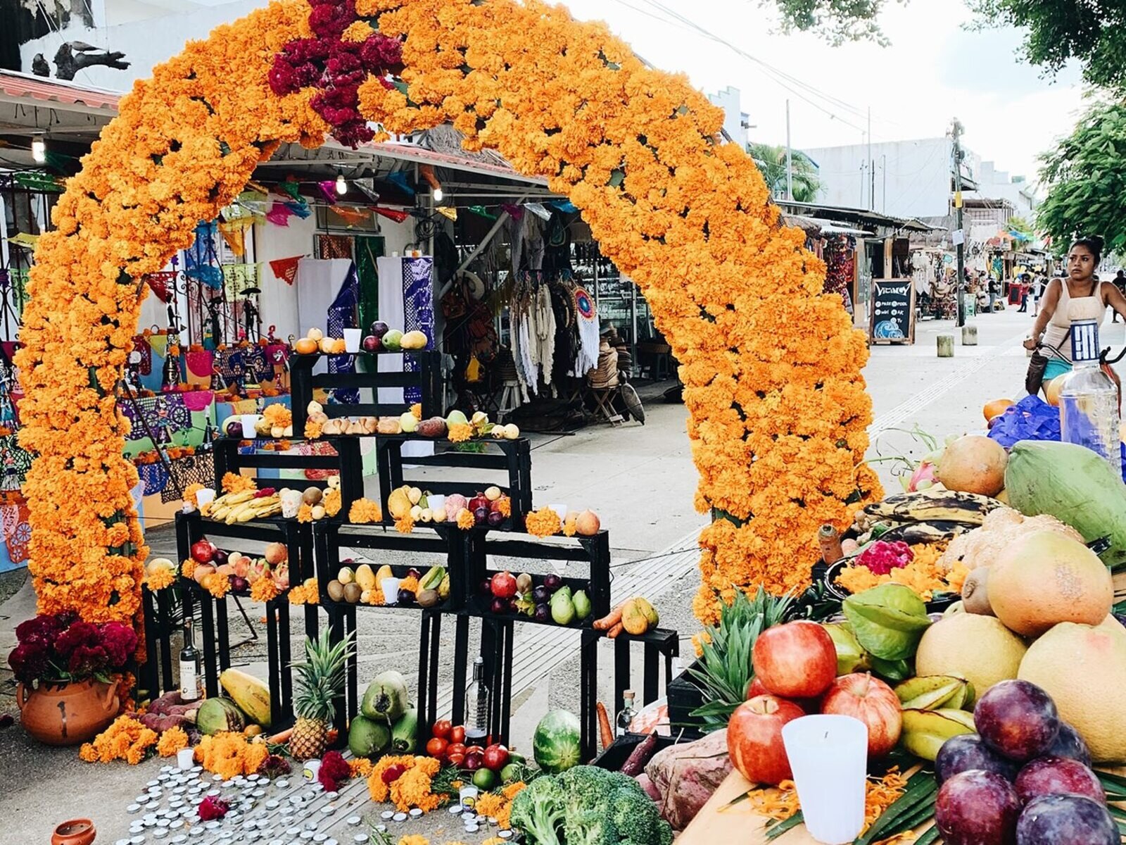Zdjęcie przedstawia łuk z kwiatów pod którym są ustawione skrzynki a na nich stoją owoce i warzywa, po prawej stoi stół pełen owoców i warzyw. W tle są stragany.