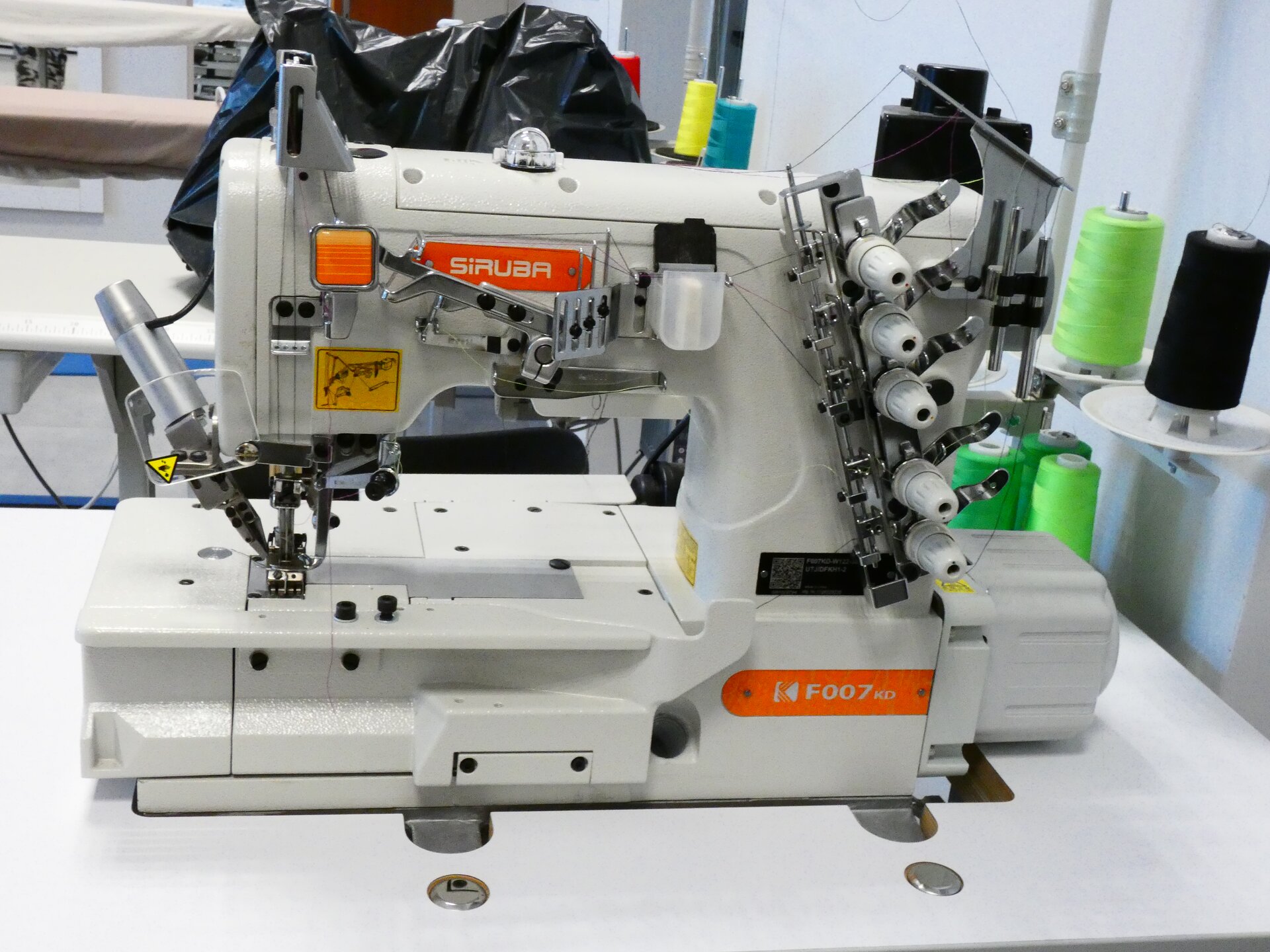 Zdjęcie przedstawia widzianą z boku białą maszynę z pomarańczowymi elementami. Składa się z ramienia z igłą oraz konstrukcji z pięcioma pokrętłami umieszczonymi jedno pod drugim.