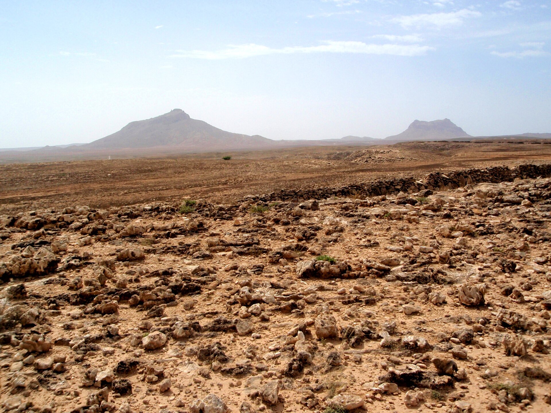 Zdjęcie przedstawia obszary pustynne na Wyspach Zielonego Przylądka. Na pierwszym planie widoczny jest piaszczysty i kamienisty, pustynny teren, a na horyzoncie dwa niewysokie wzgórza na tle błękitnego nieba.