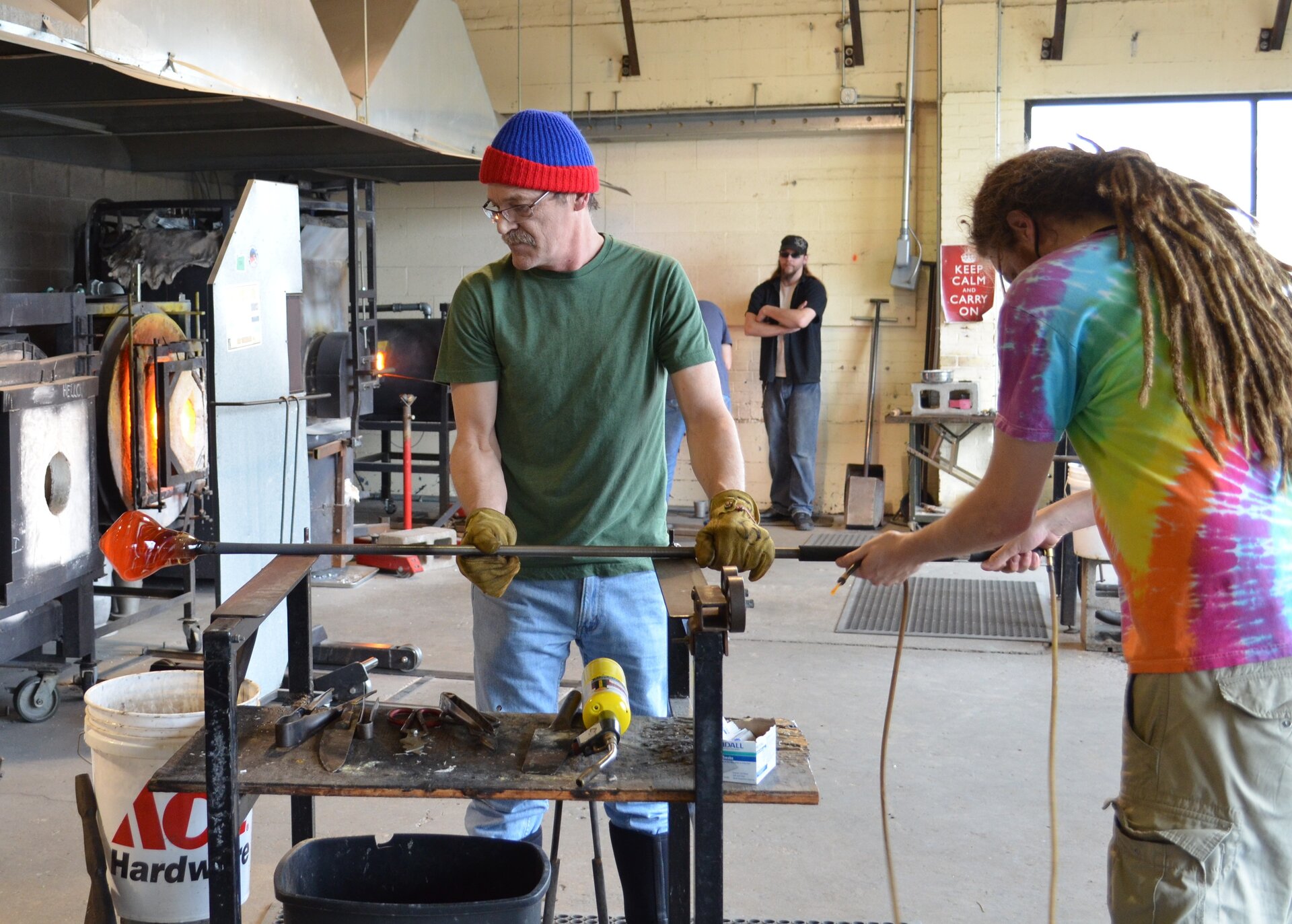 Zdjęcie koncentruje się na dwóch mężczyznach, którzy utrzymują na podstawce długi, metalowy pręt, na końcu którego znajduje się pomarańczowo‑żółta kula szkła. Jeden z nich ma na sobie rękawice ochronne. W tle piec, w którym pali się ogień. Pod ścianą pomieszczenia stoi trzeci mężczyzna.
