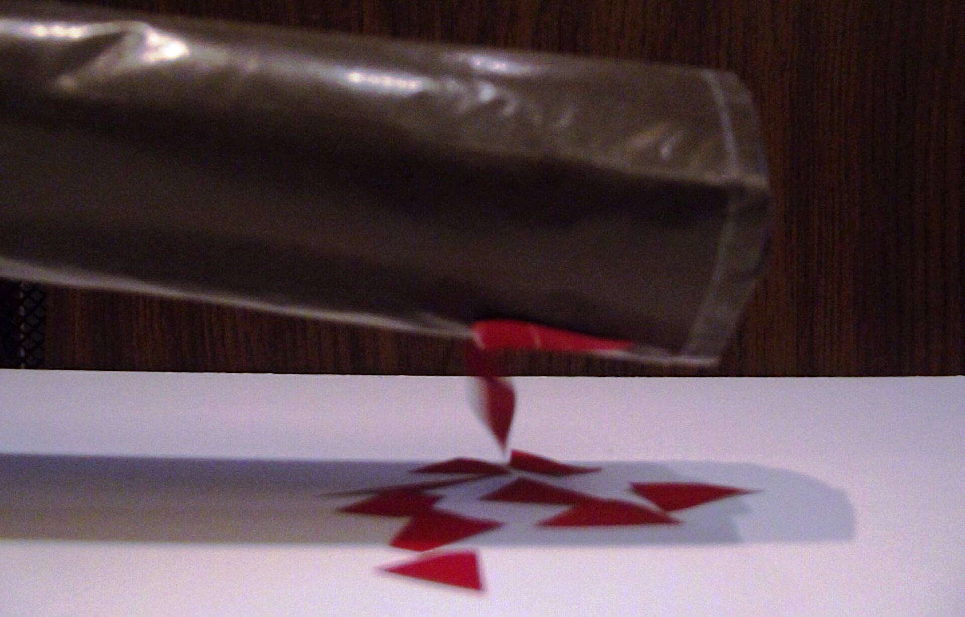 Na jasnym podłożu widać czerwone romby. Nad podłożem widać ciemnoszarą rurkę na czarnym tle. Jeden skrawek czerwony jest uniesiony nad podłożem i dotyka rurki.