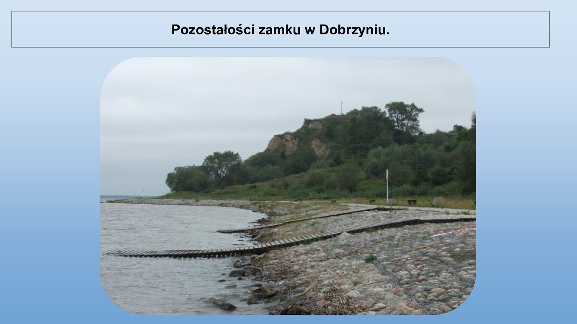 Niebieski slajd. U góry nagłówek: „Pozostałości zamku w Dobrzyniu”. Niżej zdjęcie przedstawiające brzeg wody. Z lewej strony woda, a z prawej kamienny brzeg, na którym znajdują się dwa drewniane mosty, które schodzą do wody. W tle wzgórze porośnięte zielonymi drzewami.