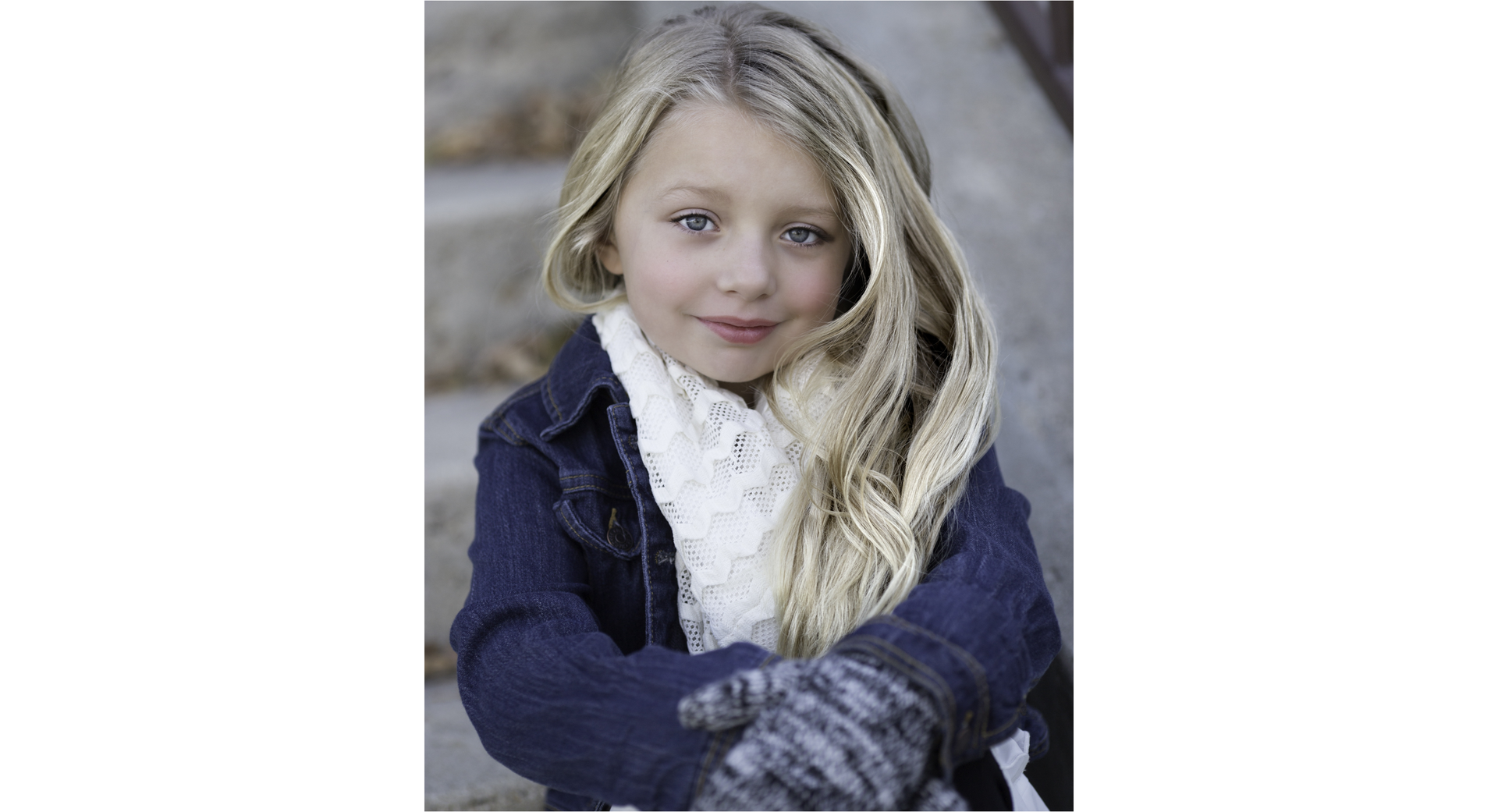 Fotografia przedstawia małą dziewczynkę z długimi jasnymi włosami. Jest ubrana w kurtkę i rękawiczki, patrzy i uśmiecha się w stronę obiektywu.