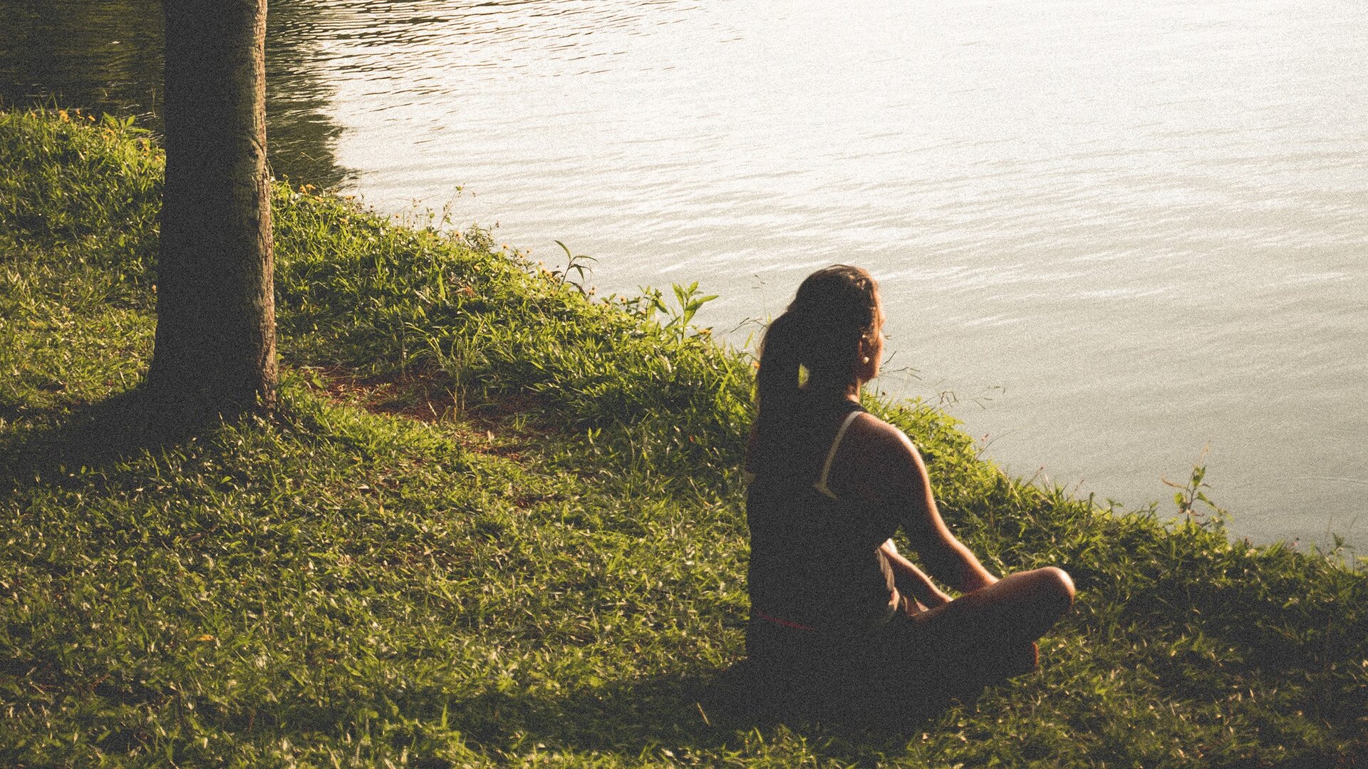 djęcie przedstawia kobietę siedzącą po turecku, widzianą od tyłu, z prawej strony. Ma długie, ciemne włosy związane w kucyk i jest ubrana w bluzkę na ramiączkach. Siedzi na trawie, nad brzegiem zbiornika wodnego, patrząc przed siebie.