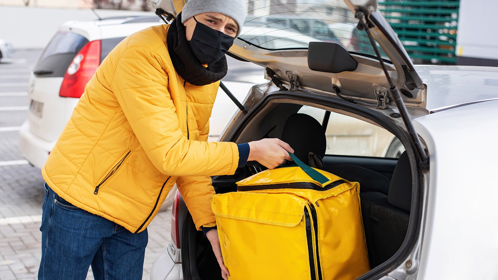 Zdjęcie przedstawia mężczyznę w maseczce medycznej, który wyjmuje z bagażnika samochodu torbę.