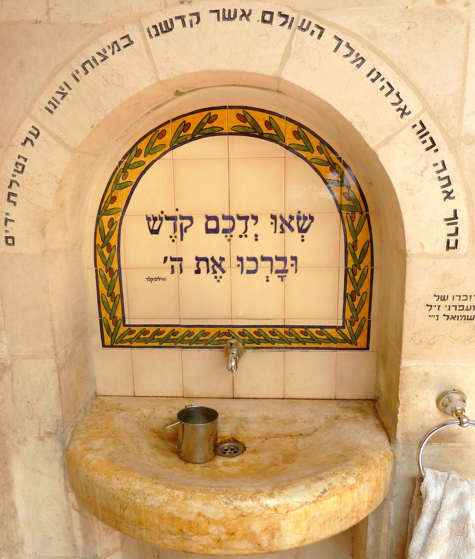 Zdjęcie przedstawia miejsce, w którym można umyć ręce i napić się z kubka wody. Po prawej stronie wisi ręcznik. Na okalającym kran łuku z kamieni są napisy w języku hebrajskim.  