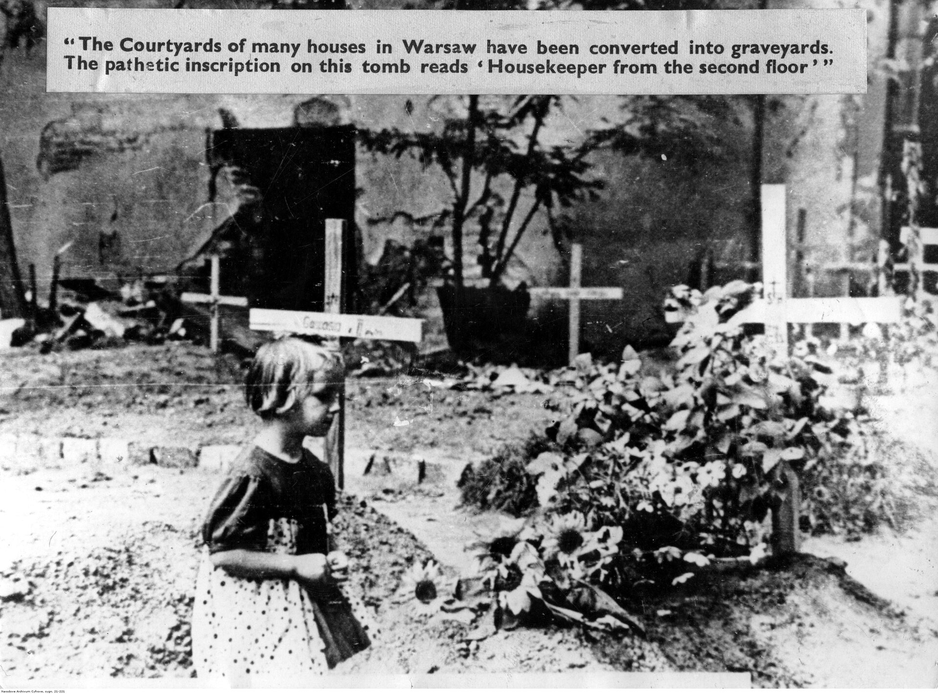 Fotografia przedstawia świeże groby z drewnianymi krzyżami między ruinami domów. Przy jednym z nich klęczy kilkuletnia dziewczynka. Dziecko ukazuje prawy profil. Ma ocienione oczy i lekko zadarty nos. Ma jasne, proste włosy sięgające za uszy. Jest ubrana w krótką sukienkę. W tle ukazane są ruiny budynku. Nad zdjęciem znajduje się napis:  The Courtyards of many houses in Warsaw have been converted into graveyards. The pathetic inscription on this tomb reads: Housekeeper from the second floor.