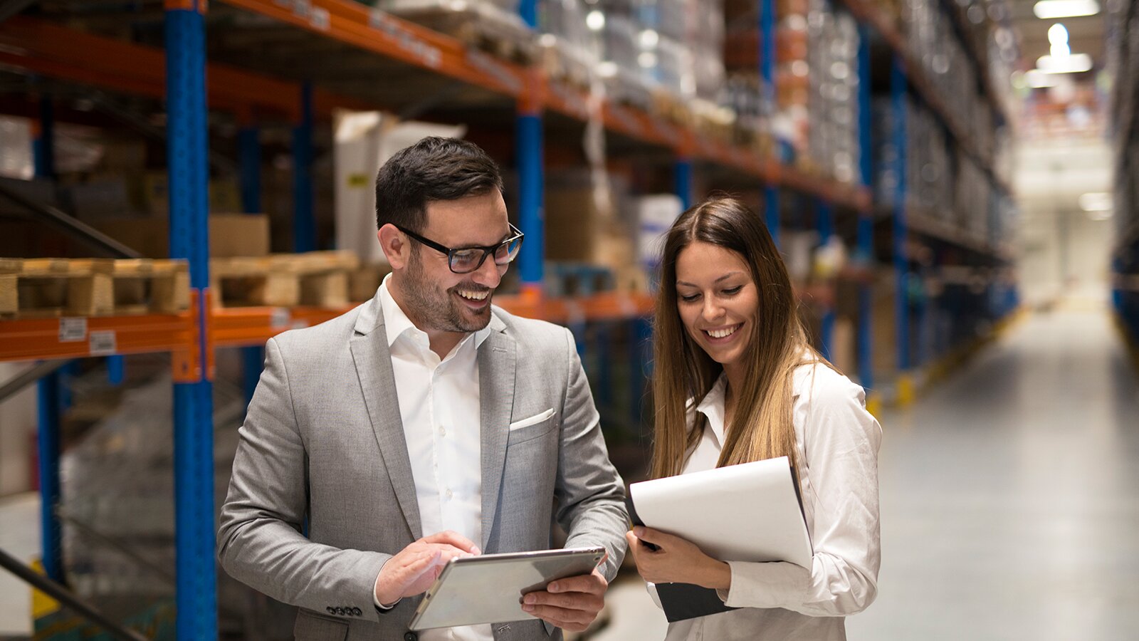 Zdjęcie przedstawia kobietę i mężczyznę stojących obok siebie w magazynie sklepowym. Oboje trzymają w dłoniach dokumenty. 