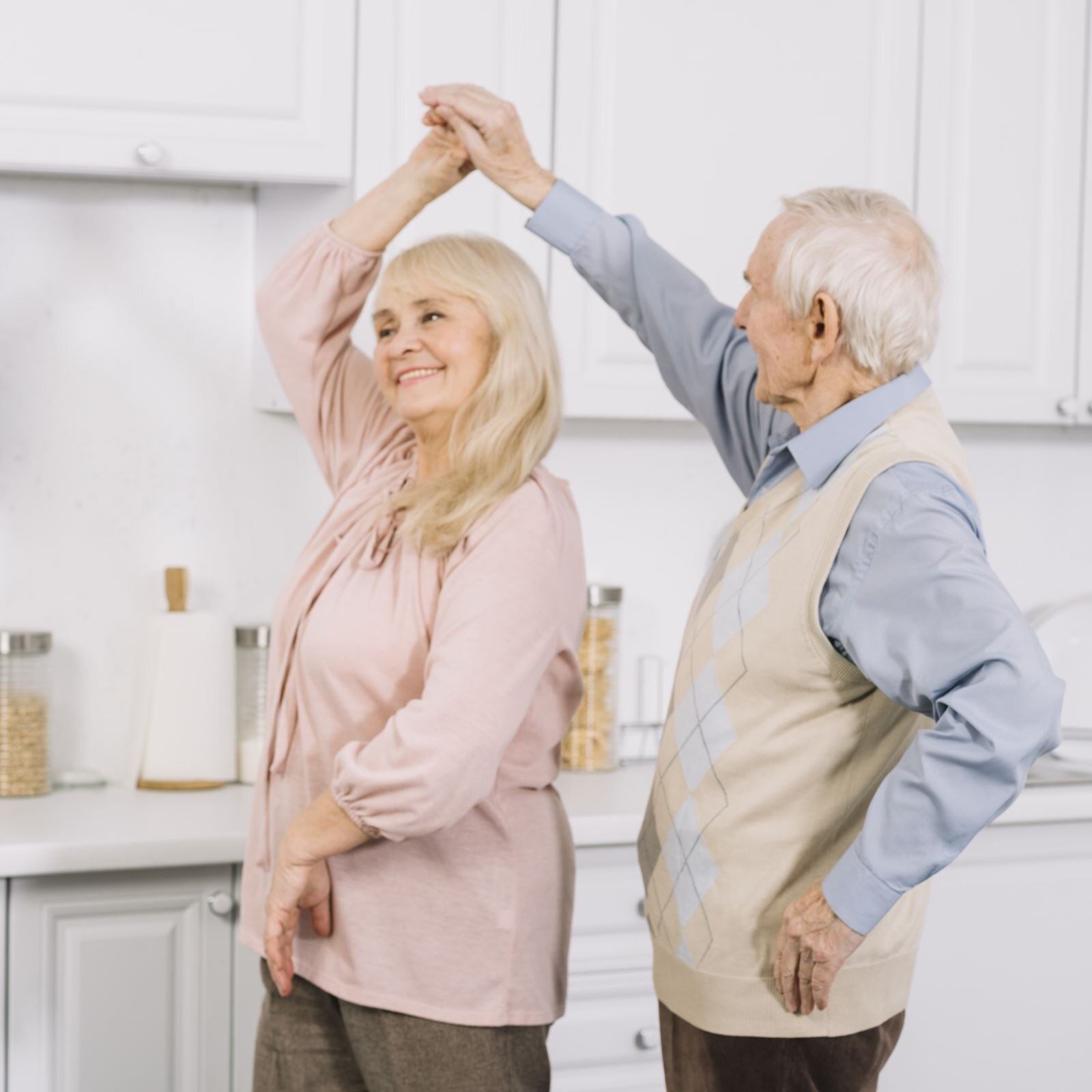 Zdjęcie przedstawia parę w wieku emerytalnym wykonującą obrót w tańcu. Kobieta ma długie, siwe włosy i jest ubrana w różową bluzkę. Prawą rękę ma podniesioną nad głową, a lewą, zgiętą w łokciu, opartą na brzuchu. Mężczyzna stojący po prawej ma krótkie siwe włosy i jest ubrany w niebieską koszulę oraz kremową kamizelkę z rombami na przodzie. Prawą ręką trzyma prawą dłoń kobiety, a lewą, zgiętą w łokciu, ma opartą na biodrze. W tle widoczna jest kuchnia z białymi meblami.