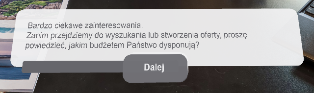 Zdjęcie przedstawia widok ekranu z poleceniem. Na tle biurka widoczny jest biały prostokąt w poziomie z zaokrąglonymi narożnikami, na którym znajdują się szare napisy, pośrodku którego znajduje się pole z napisem Dalej.