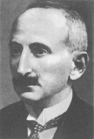 Kliknij, aby powiększyć. Zdjęcie przedstawia portret mężczyzny z półprofilu o krótko przystrzyżonych, siwiejących włosach i czarnych wąsach. Ma na sobie białą koszulę ze sztywnym kołnierzykiem, krawat i ciemną marynarkę.