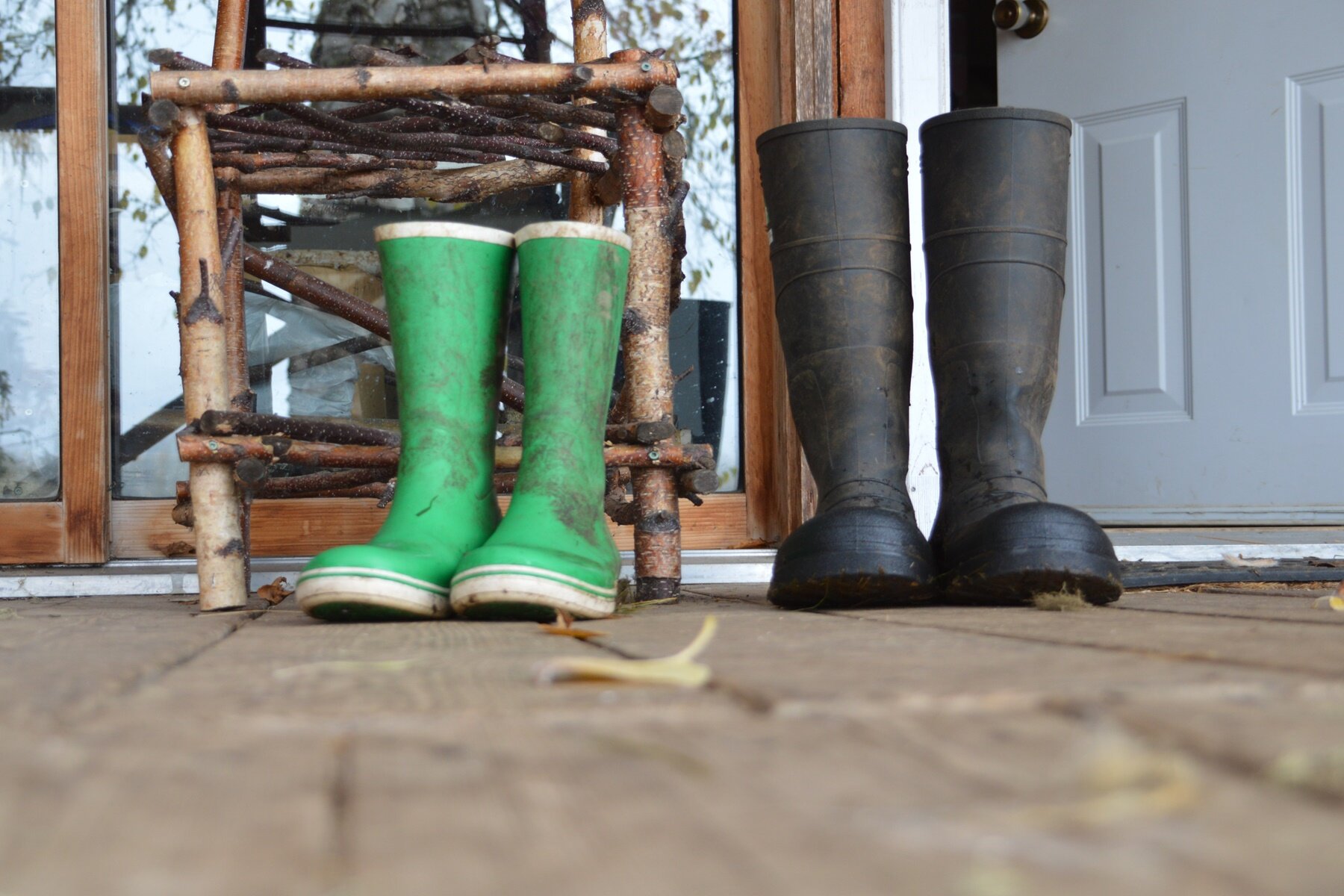 Zdjęcie przedstawia dwie pary brudnych kaloszy, stojących na drewnianej podłodze przed wejściem. Po lewej stronie widać mniejsze kalosze w kolorze zielonym, a po prawej stronie większe w kolorze czarnym. W tle widoczna szafla z drewna, za którą widać szyby w drewnianych ramach. Na prawo od nich widać otwarte białe drzwi.