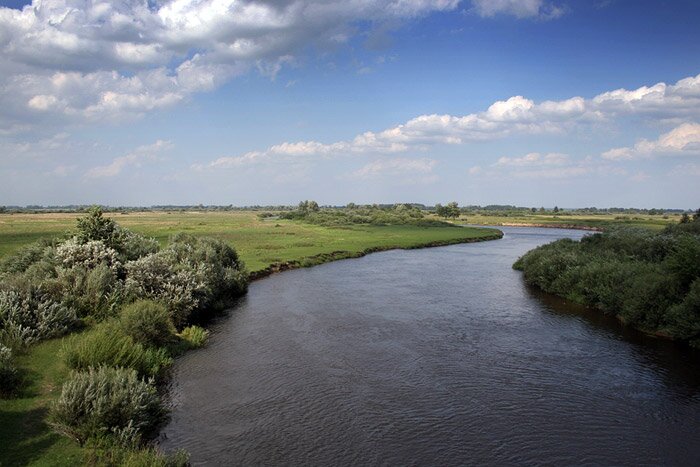 Na zdjęciu spokojny, sielski krajobraz. Na pierwszym planie znajduje się rzeka o szerokim korycie, w tle jej koryto zakręca w prawą stronę. Po lewej i prawej stronie rzeki znajdują się rozległe tereny zielone: pola, zarośla i drzewa. Nad rzeką znajduje się błękitne niebo, częściowo pokryte białymi, lekkimi chmurami. 