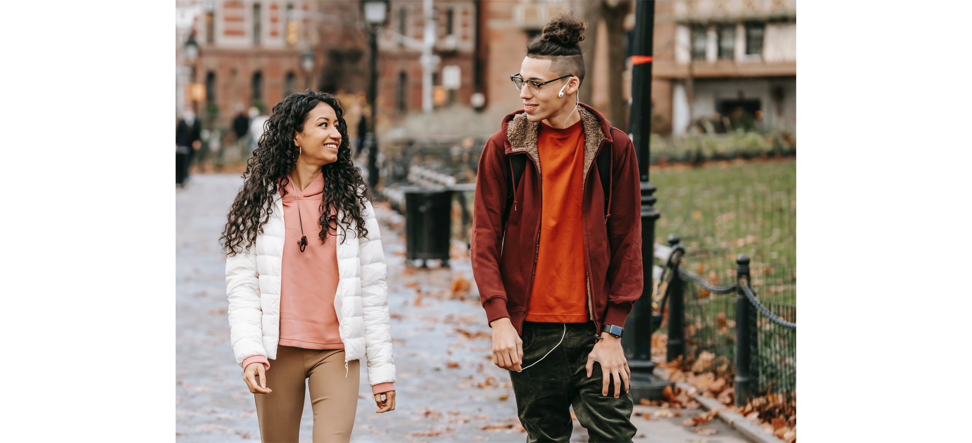 Zdjęcie przedstawia dziewczynę i chłopaka idących ulicą. Dziewczyna znajduje się po lewej stronie zdjęcia, ma długie kręcone włosy, jest uśmiechnięta. Po prawej stronie znajduje się chłopak w okularach, ma włosy zaczesane do góry w kitkę.