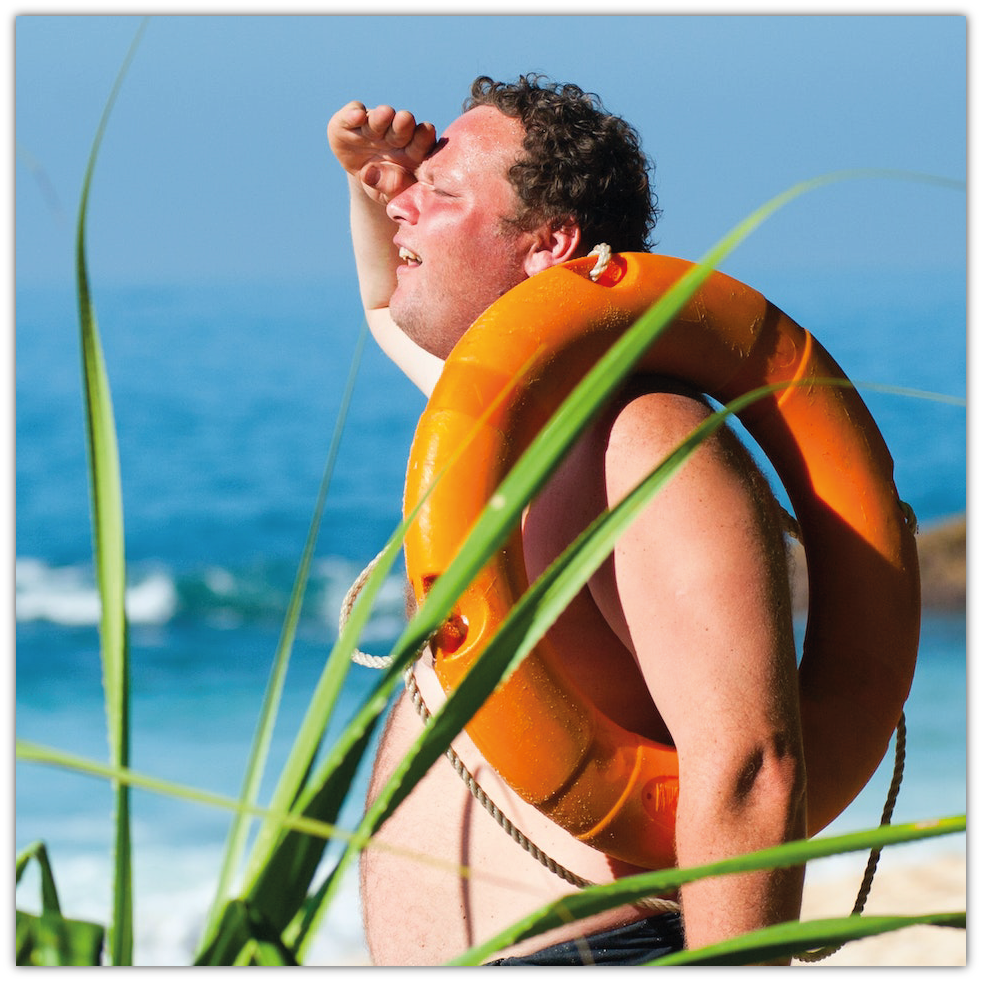 Zdjęcie przedstawia mężczyznę w średnim wieku stojącego na plaży. Mężczyzna nie ma na sobie koszulki, przez ramię przełożył sobie pomarańczowe koło ratunkowe, a dłoń przykłada do czoła patrząc w dal.