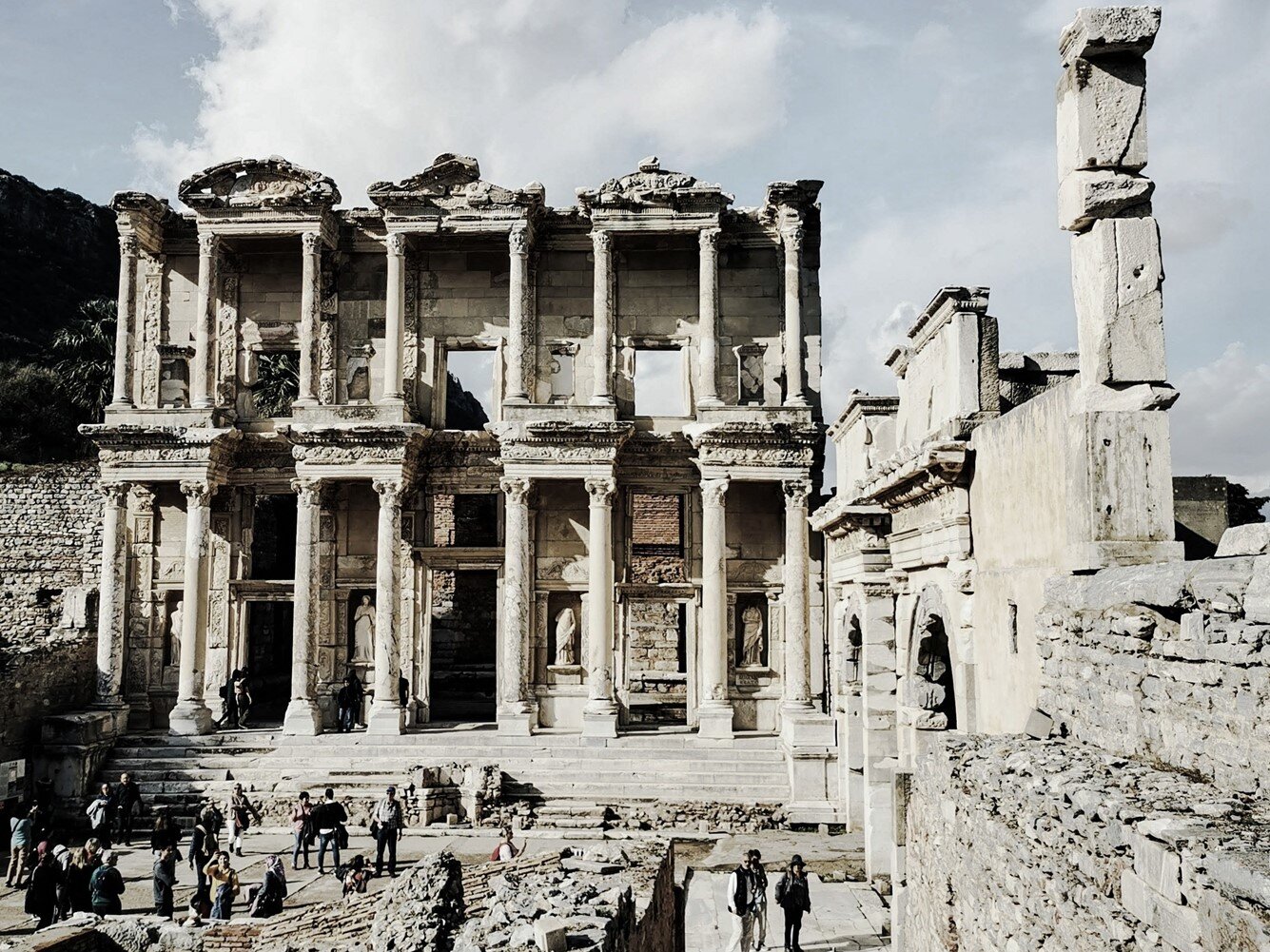 Zdjęcie przedstawia antyczną budowlę. Widoczny jest okazały budynek składający się z dwóch kondygnacji. Do budynku prowadzą schody. Na każdej z kondygnacji widoczne są cztery pary kolumn. Na parterze pomiędzy kolumnami znajdują się wejścia oraz nisze z posągami. Na piętrze widoczne są balkony, a pary kolumn połączone są trójkątnymi tympanonami. Po prawej stronie widoczny jest antyczny mur z dwoma łukami. Przed budynkiem widoczne są grupy ludzi.