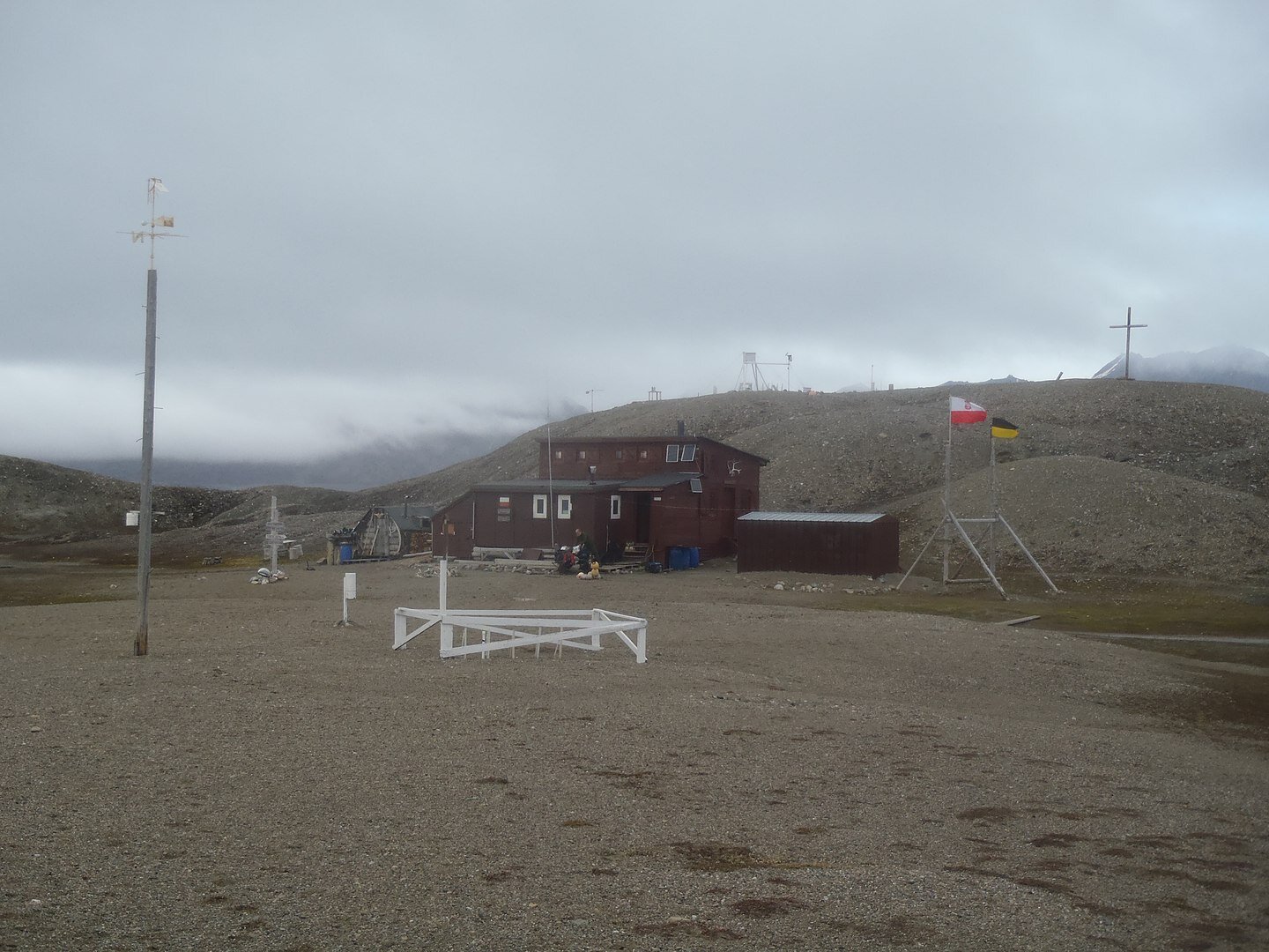 Zdjęcie przedstawia niewielki budynek stacji badawczej o długości kilku metrów. Przed nim płaski teren, za nim wzniesienia. Całość pokryta drobnymi kamykami.