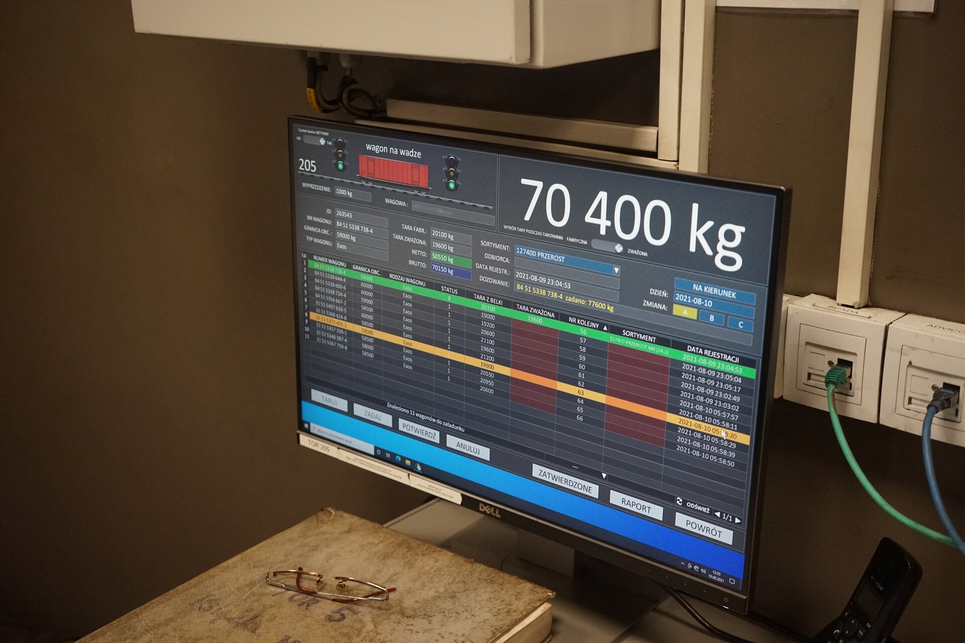 Na zdjęciu widoczne jest stanowisko operatora nasypywarki. W centralnej części zdjęcia znajduje się ekran komputera. Na ekranie w prawym górnym rogu wyświetla się wartość: siedemdziesiąt tysięcy czterysta kilogramów. Niżej widoczne są tabele z danymi.Poniżej zdjęcia widoczny jest przycisk multimedialny z nagraniem audio. Treść nagrania jest zgodna z tytułem zdjęcia.