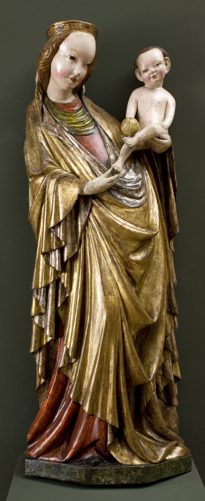 Rzeźba przedstawia Matkę Boską z Dzieciątkiem. Kobieta ma na sobie obszerne, złote szaty aż do ziemi, spod których wystaje czerwony materiał. Ma ona pulchną twarz z okrągłymi policzkami, dużym czołem, drobnymi ustami, dużymi oczami i cienkimi brwiami. Twarz ma zwrócona w stronę dziecka. Na głowie ma lekko falowane, długie włosy. Na głowie ma złotą, okrągłą koronę z namalowanymi detalami. Jej ciało jest odchylone po skosie do tyłu, a na swojej lewej ręce trzyma małego chłopca. Chłopiec siedzi wyprostowany, z głową przechyloną w prawą stronę, wzrok patrzy w przód. Ma okrągłą twarz, pulchne policzki, duże oczy, duże uszy. Na głowie ma brązowe włosy. W prawej ręce trzyma złote, okrągłe jabłko. Prawą stopę wyciąga w dół, a kobieta trzyma go za stopę prawą dłonią. Rzeźba ustawiona jest na wielobocznej podstawie. Tło jest ciemnoszare.