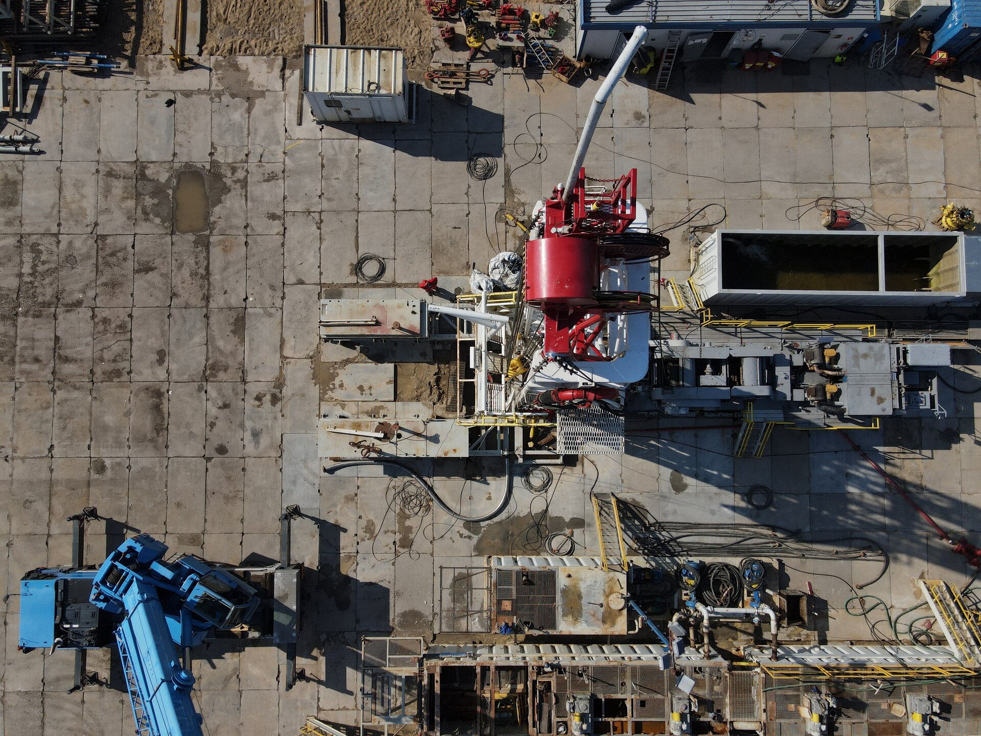 Na zdjęciu widać maszt urządzenia wiertniczego widziany z loku ptaka. Widoczny na zdjęciu maszt znajduje się na betonowym placu. Maszt jest konstrukcją kratową o kształcie prostopadłościanu o orientacji pionowej. W dolnej części zdjęcia widoczny jest podest, który jest w trakcie budowy. Na górze wieży widać walec. Po walcu biegnie w dół stalowa lina. Jest to wielokrążek. W otoczeniu masztu widać zbiorniki. 