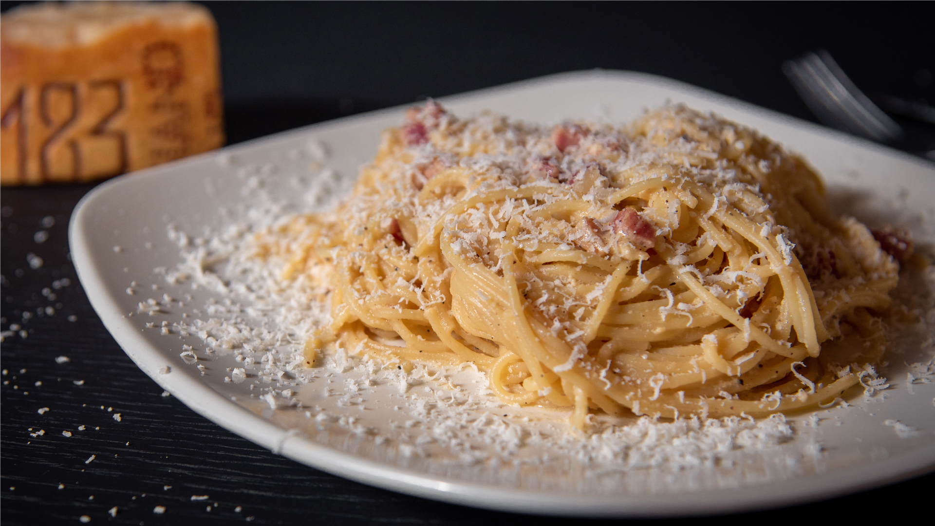 Zdjęcie przedstawia gotowe danie spaghetti alla carbonara na talerzu. Nitki makaronu ułożone są w kształcie gniazda. Widoczne są kawałki smażonego boczku. Danie posypane jest serem.
