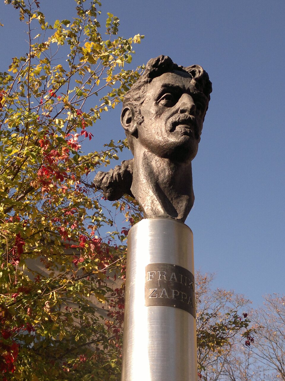 Ilustracja przedstawia rzeźbę Konstantinasa Bogdanasa „Pomnik Franka Zappy”. Ukazuje uniesioną głowę artysty, ustawioną na wysokim, metalowym słupie z inskrypcją.