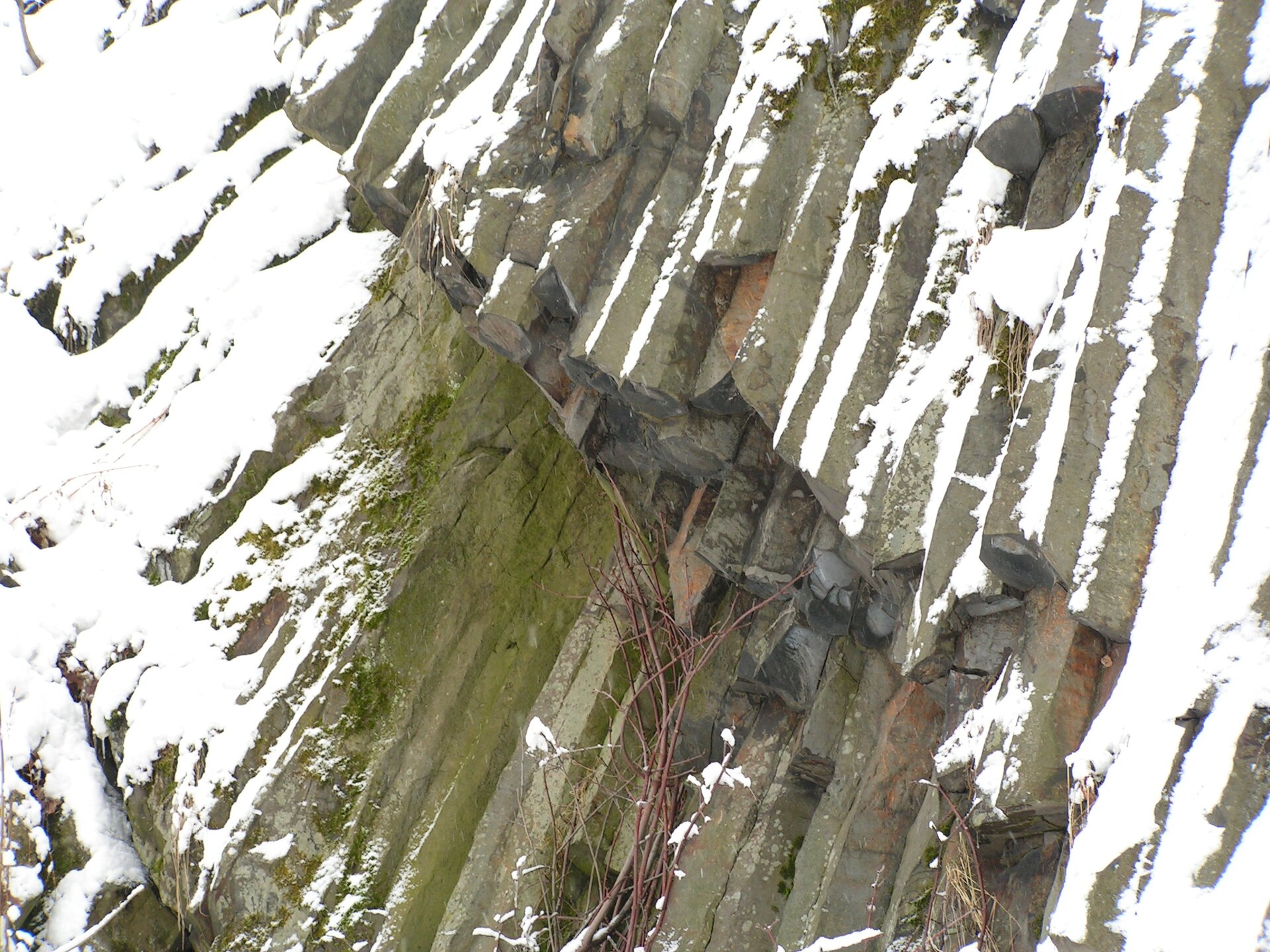 Na zdjęciu bazaltowe wielokątne w przekroju słupy skalne na zboczu pokrytym częściowo śniegiem.