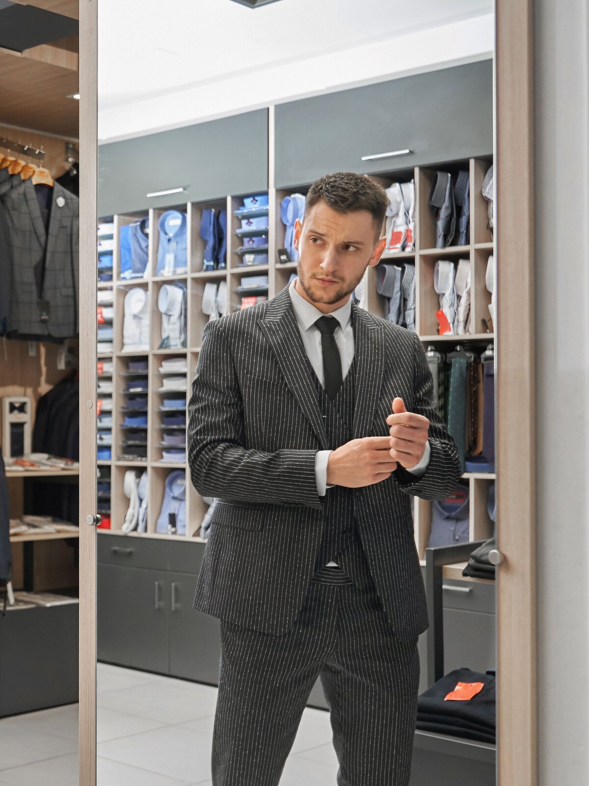 Zdjęcie przedstawia młodego mężczyznę ubranego w szary, prążkowany garnitur oraz białą koszulę, krawat i prążkowaną kamizelkę. 