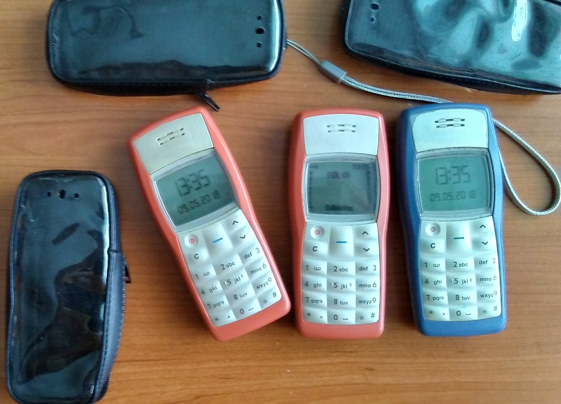  Zdjęcie przedstawia drewniany blat, na którym leżą trzy stare modele telefonów komórkowych, z dużymi klawiaturami numerycznymi i przyciskami oraz bardzo małymi wyświetlaczami. Dwa telefony z prawej mają jasnoczerwoną obudowę, a trzeci obudowę w kolorze niebieskim. Po lewej stronie i powyżej telefonów widoczne są trzy czarne etui na telefony. 