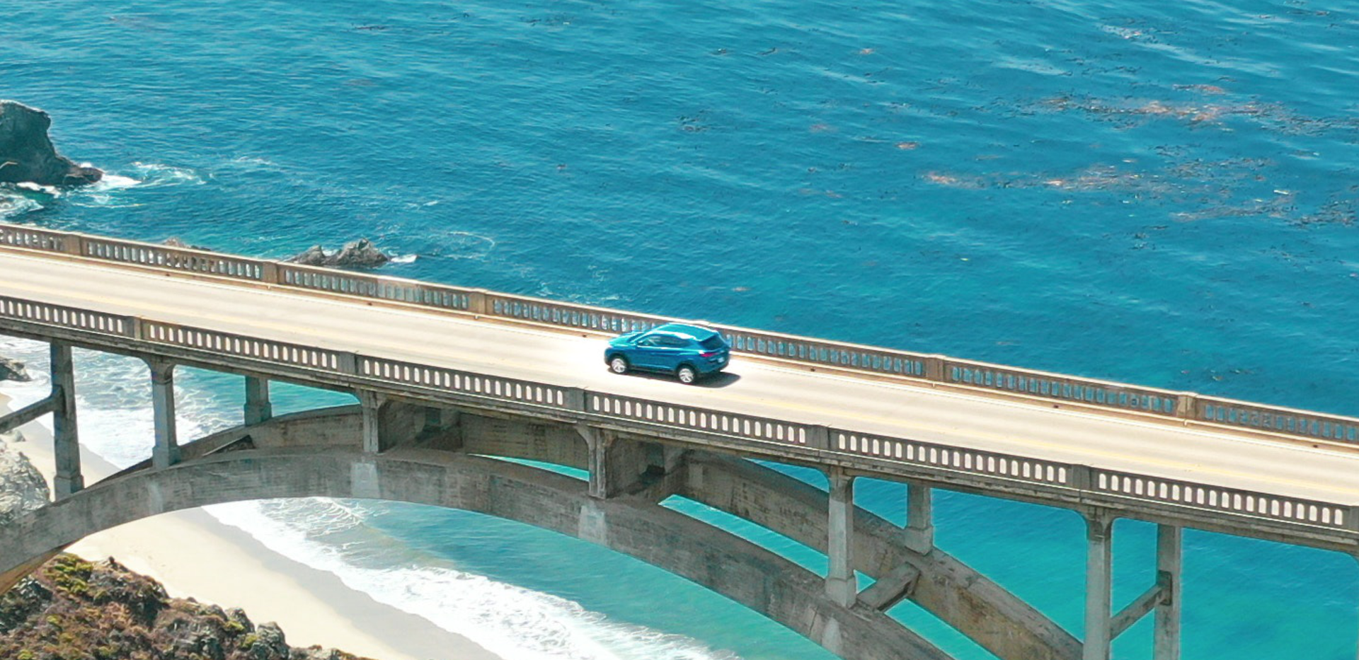 Zdjęcie wykonane z góry przedstawia samochód osobowy przejeżdżający mostem. Samochód przejeżdża nad lazurowym morzem. Jest piękna pogoda, świeci słońce.