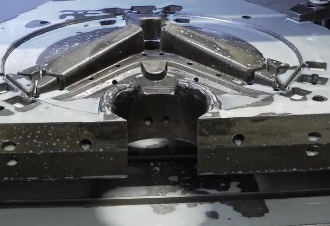 Ilustracja przedstawia fragment metalowej formy odlewniczej. Widoczny jest obszar układu wlewowego z napawanym metalem. Połówka formy ustawiona jest na stoliku roboczym frezarki.