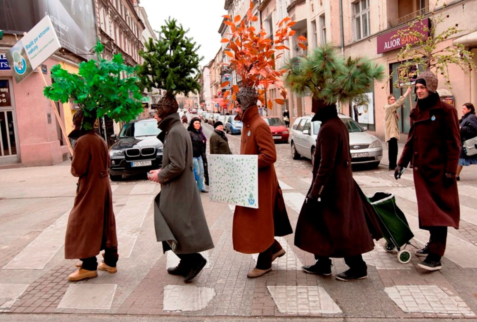 Zdjęcie przedstawia młodych mężczyzn przechodzących rzędem przez ulicę Żywca‑Zdroju.  Mężczyźni ubrani są w długie płaszcze , na czapkach zamontowane mają stylizowane gałęzie drzew liściastych i iglastych, niosą planszę: "Bądź po stronie natury". To happening ekologiczny, który namawia w ten sposób do ochrony przyrody.
