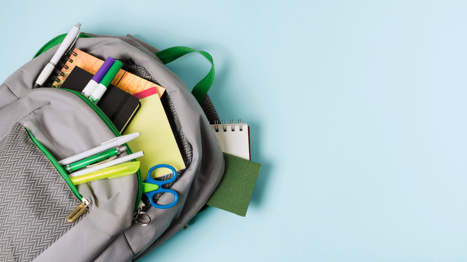 Zdjęcie przedstawia plecak szkolny, z którego wystają różne przybory. Widoczne są: zeszyty, notatniki, długopisy, ołówki, markery, nożyczki oraz ekierka.
