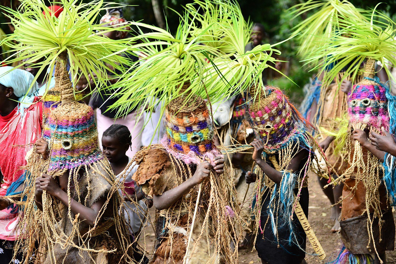 Fotografia. Na niej czworo czarnoskórych ludzi, przebranych w lniane i skórzane ubrania. na głowie mają kolorowe maski ze sznurka. Na czubku wykończenie z trawy przypominające palmę. Osoby te są w ruchu. Za nimi inni ludzie. 
