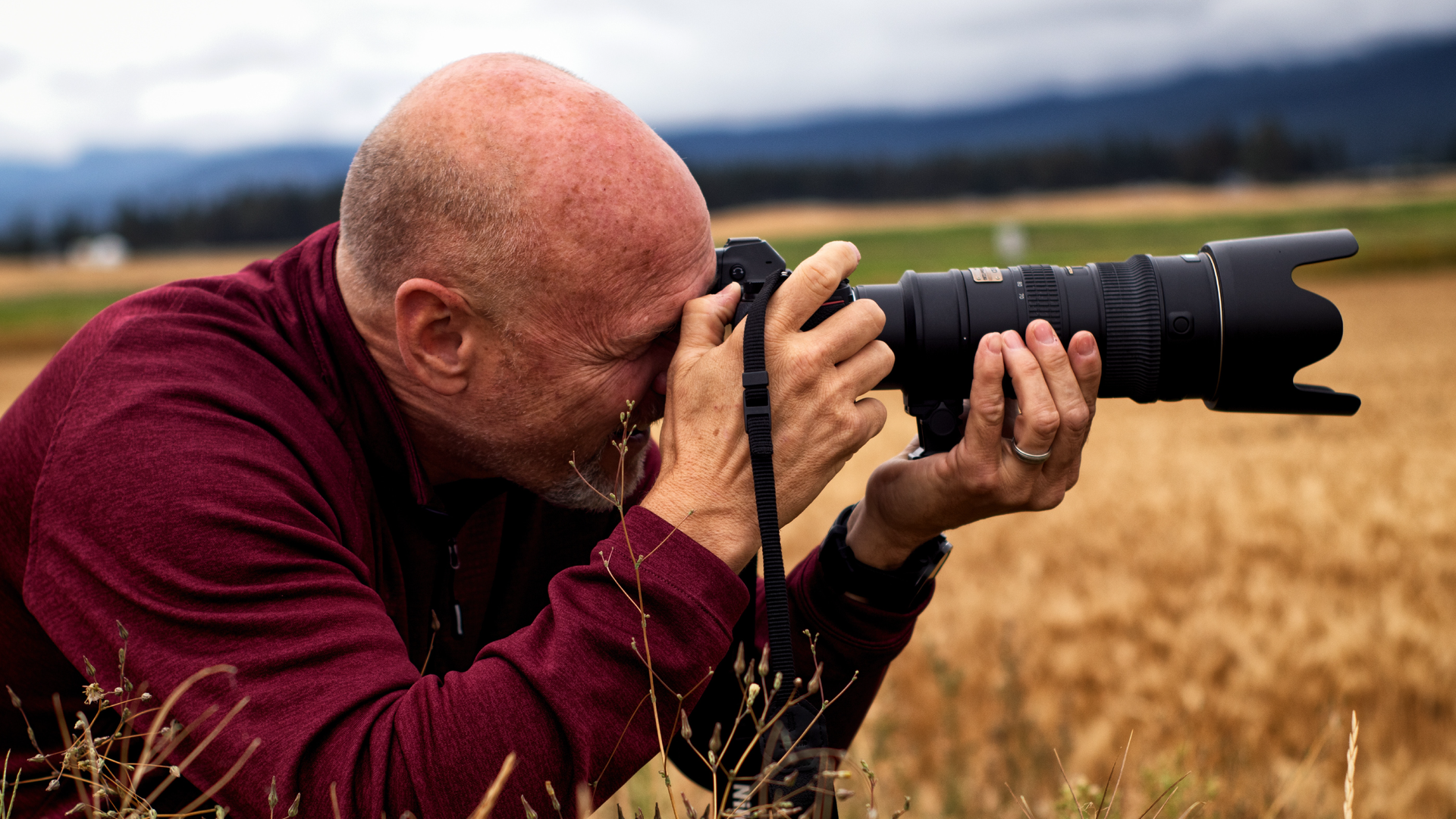 Zdjęcie przedstawia zbliżenie na łysego mężczyznę, który trzyma przy twarzy aparat fotograficzny. Mężczyzna robi zdjęcie. Aparat fotograficzny na bardzo długi obiektyw.
