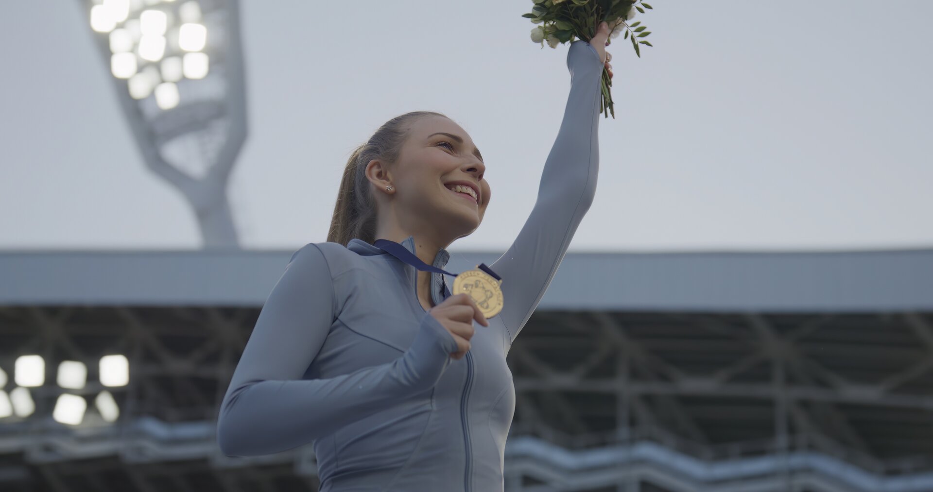 Ilustracja przedstawia uśmiechniętą kobietę w sportowym stroju, która w jednej dłoni trzyma złoty medal, drugą, trzymając bukiet kwiatów, unosi do góry.  W tle widać oświetlenie hali sportowej. 