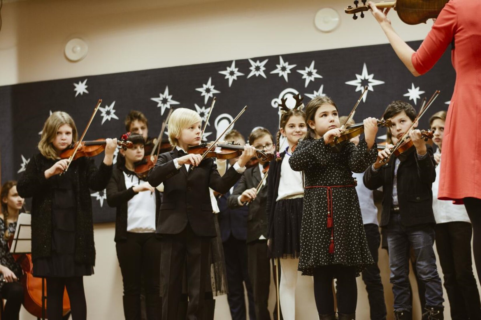 Fotografia z koncertu dziecięcego z Częstochowskiej Szkoły Suzuki, przedstawia grupkę dzieci w wieku szkoły podstawowej, które ćwiczą granie na skrzypcach. Naprzeciwko nich stojąca przodem nauczycielka, pokazuje dzieciom jak trzymać skrzypce. Każde dziecko ma prawidłowo ułożone dłonie do grania na skrzypcach. Lewe dłonie mają ułożone na gryfie ze strunami, a w prawej trzymają smyczek muskając je. Dzieci ubrane są na galowo, natomiast nauczycielka w czerwoną sukienkę do kolan. Jedna dziewczynka ma na głowie opaskę z rogami renifera. Na drugim planie po lewej stronie znajduje się siedząca dziewczynka z opartym o nogi dużym, drewnianym instrumentem muzycznym. Dziewczynka ma przed sobą białe kartki papieru, ułożone na czarnym stojaku. W tle na beżowej ścianie widać czarną, dużą tablicę ze sporą ilością przyklejonych, białych gwiazdek. 