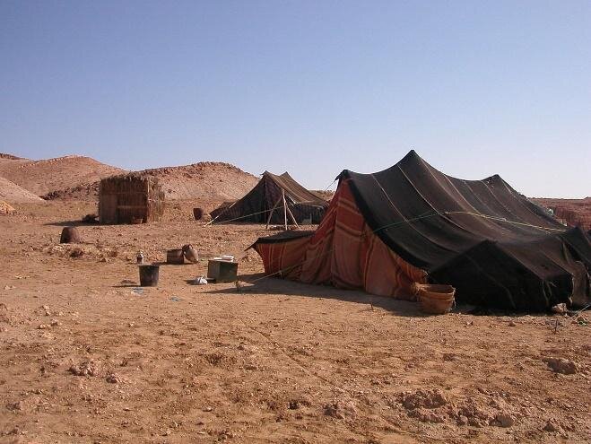 Fotografia przedstawiająca czarne namioty rozłożone na pustyni. Przed nimi pojedyncze przedmioty, takie jak wiadra czy kosze. Dookoła rozległy, pustynny, płaski teren, brak roślinności.