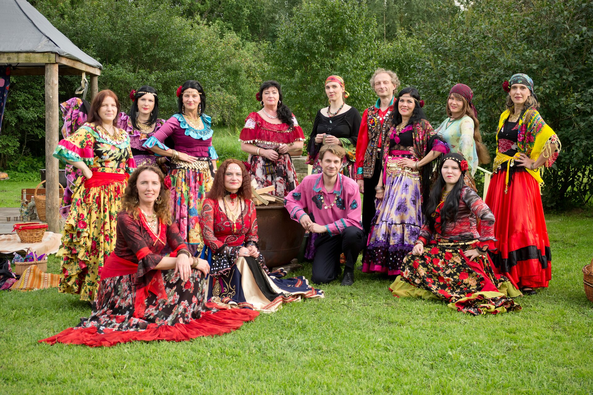 Na zdjęciu grupa kilkunastu kobiet romskich w bardzo kolorowych, zdobionych, rozłożystych sukniach. Stoją na trawie, obok altana, na drugim planie drzewa i krzewy. Pośród kobiet dwóch mężczyzn, noszą luźne, kolorowo zdobione koszule.