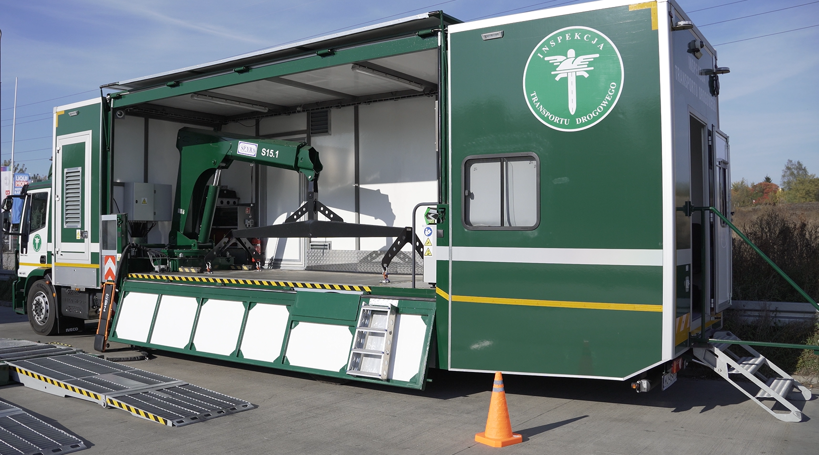 Zdjęcie przedstawia specjalistyczny samochód inspektoratu transportu drogowego. Samochód jest ciemno zielony. Wewnątrz samochodu znajduje się specjalny dźwig, dzięki któremu możliwe jest składanie i rozkładanie wagi. Przed samochodem rozłożona jest waga służąca do ważenie pojazdów ciężarowych. 