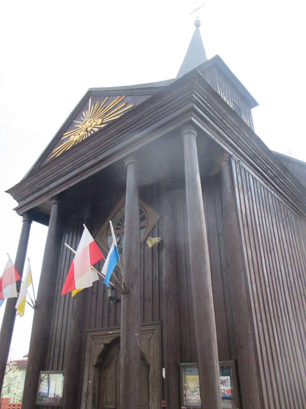 Zdjęcie przedstawia wejście do drewnianego kościoła. Nad wejściem znajduje się daszek, na którym znajdują się złote zdobienia. Daszek przytrzymywany jest drewnianymi kolumnami. Nad daszkiem widoczny jest fragment wieżyczki zakończonej skośnym dachem, na czubku którego znajduje się krzyż. Przy wejściu umieszczone zostały trzy flagi: biało‑czerwona, biało‑żółta oraz biało‑niebieska.